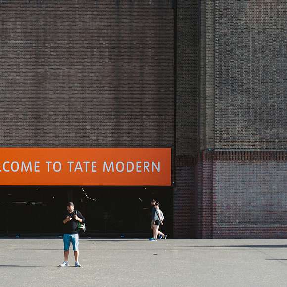 Exterior of Tate Modern with an orange sign reading “Welcome to Tate Modern” and visitors walking outside.