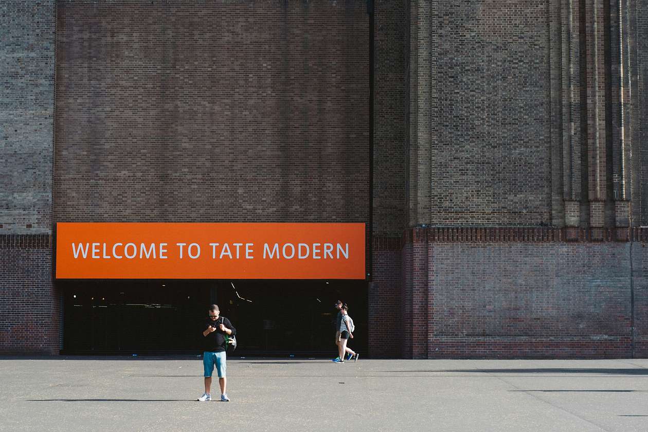 Exterior of Tate Modern with an orange sign reading “Welcome to Tate Modern” and visitors walking outside.