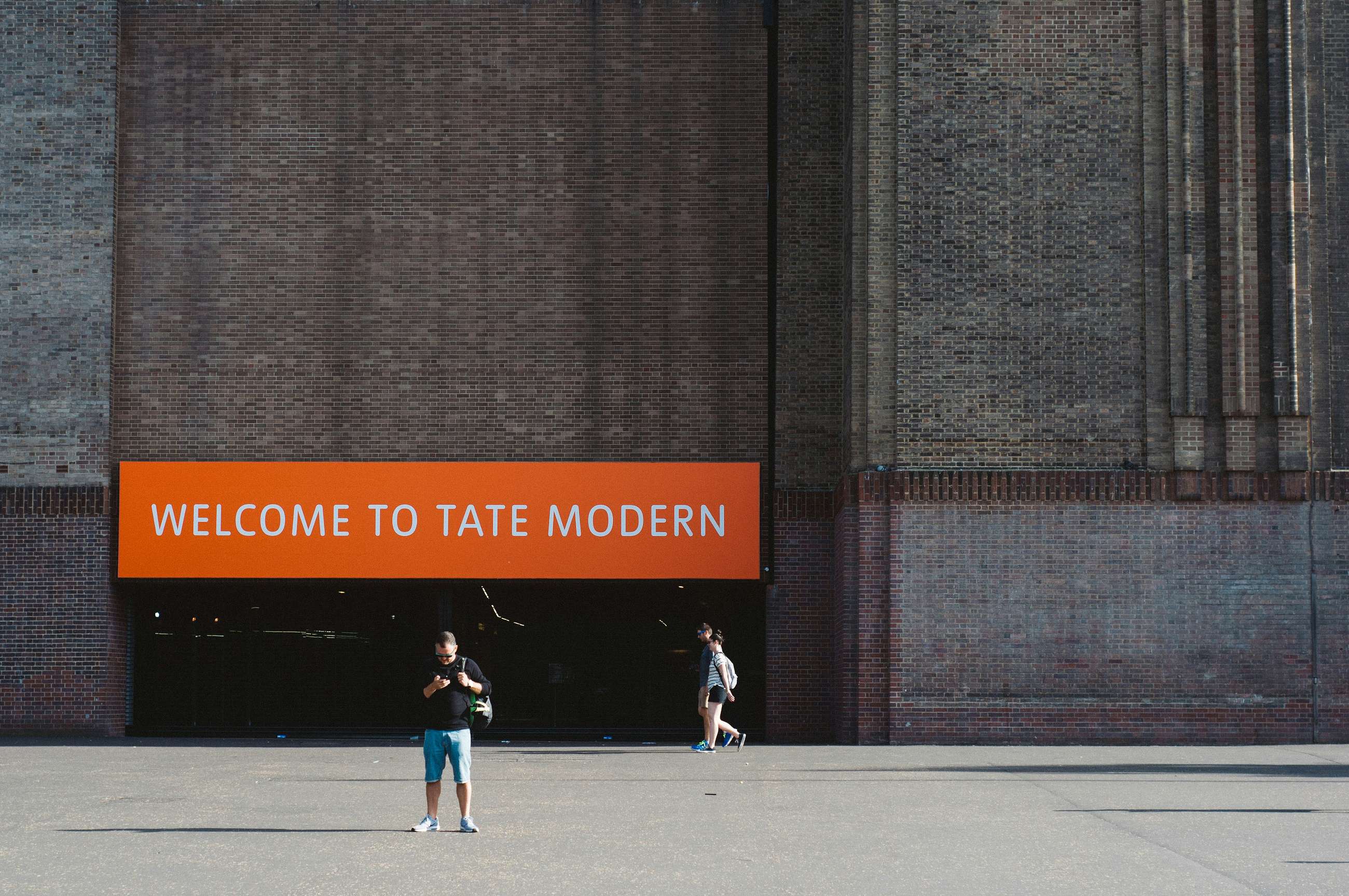 Exterior of Tate Modern with an orange sign reading “Welcome to Tate Modern” and visitors walking outside.