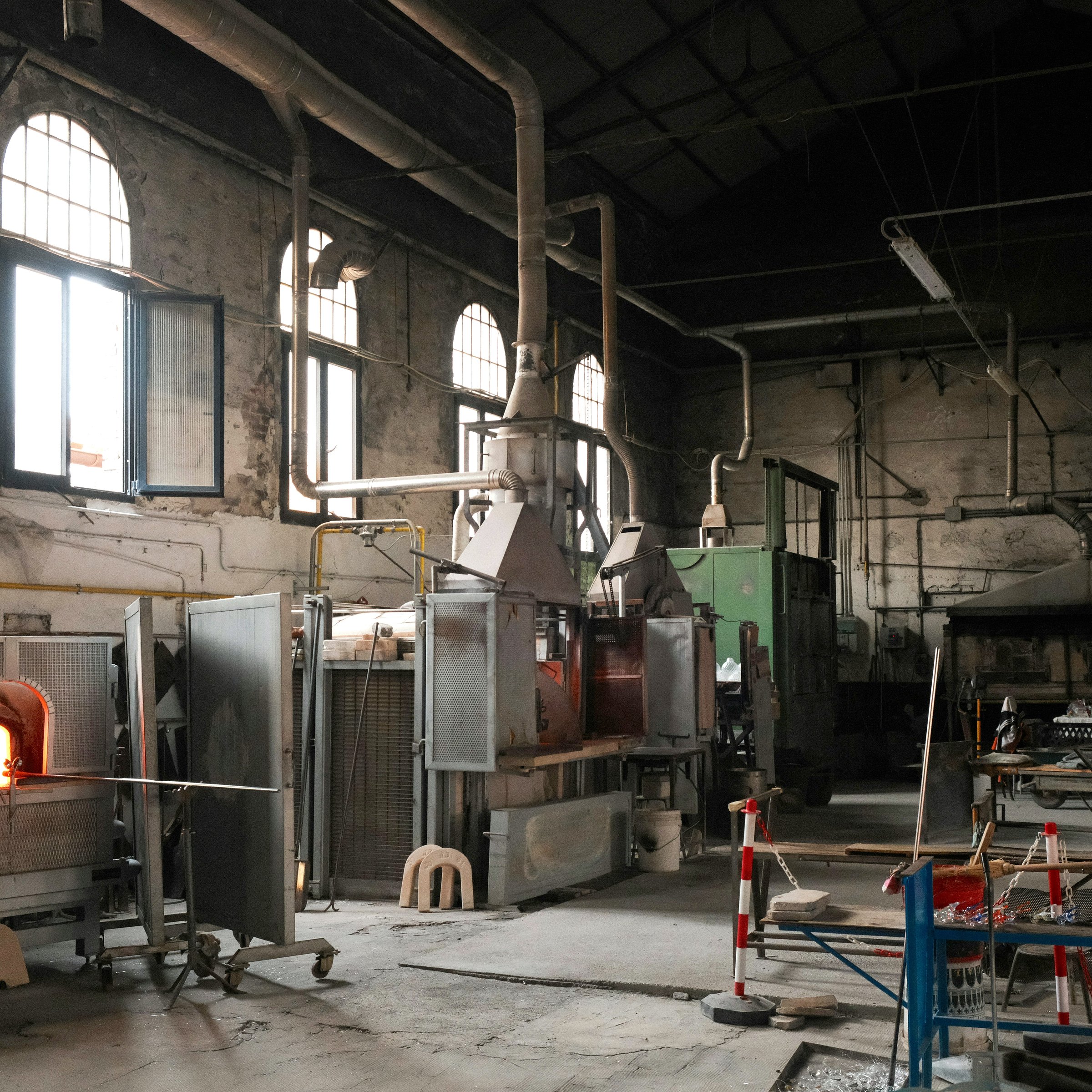 An interior view of a traditional glassblowing workshop with large furnaces, ventilation pipes, and scattered tools and materials throughout the space.