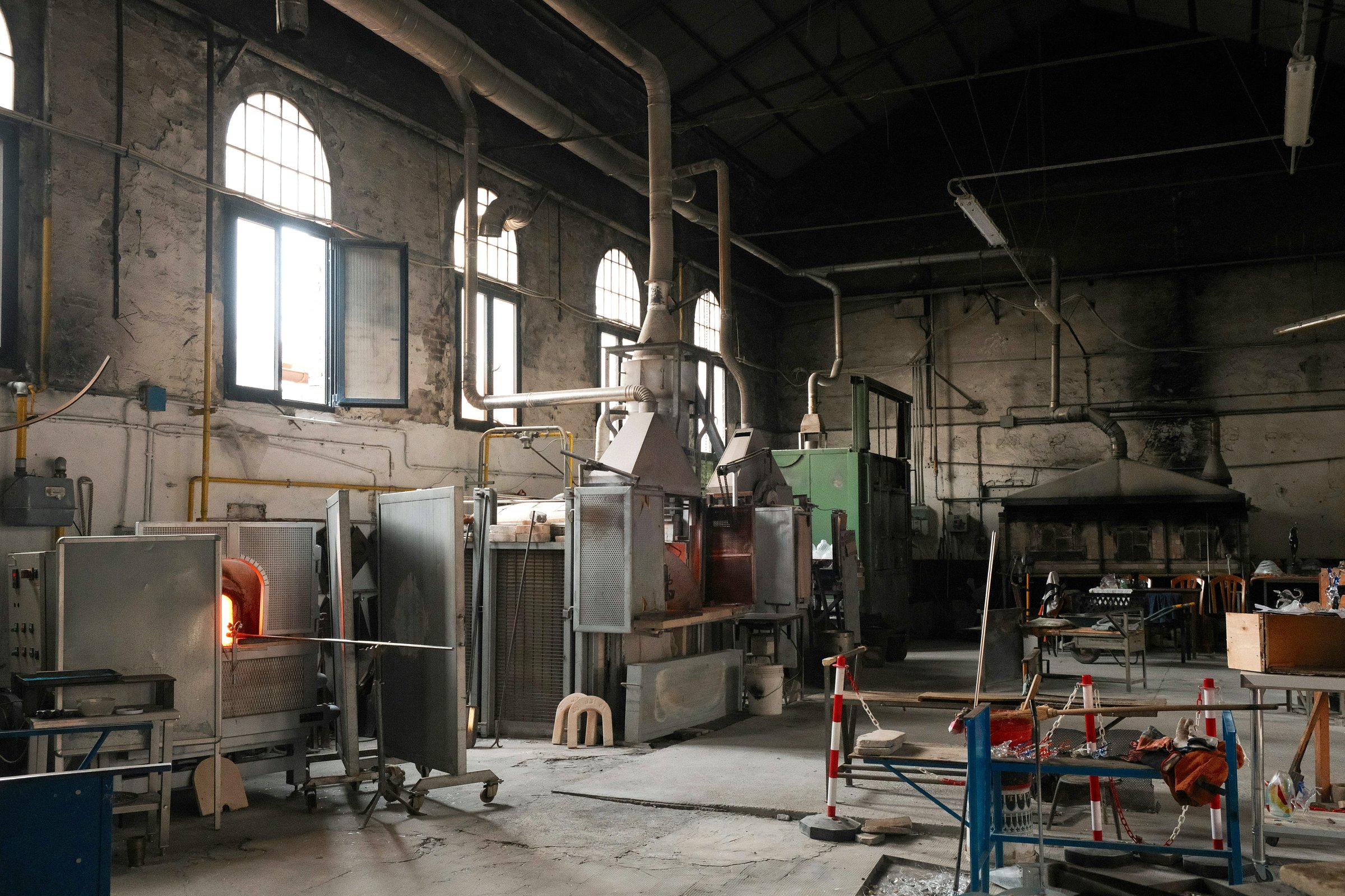 An interior view of a traditional glassblowing workshop with large furnaces, ventilation pipes, and scattered tools and materials throughout the space.
