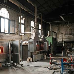 An interior view of a traditional glassblowing workshop with large furnaces, ventilation pipes, and scattered tools and materials throughout the space.