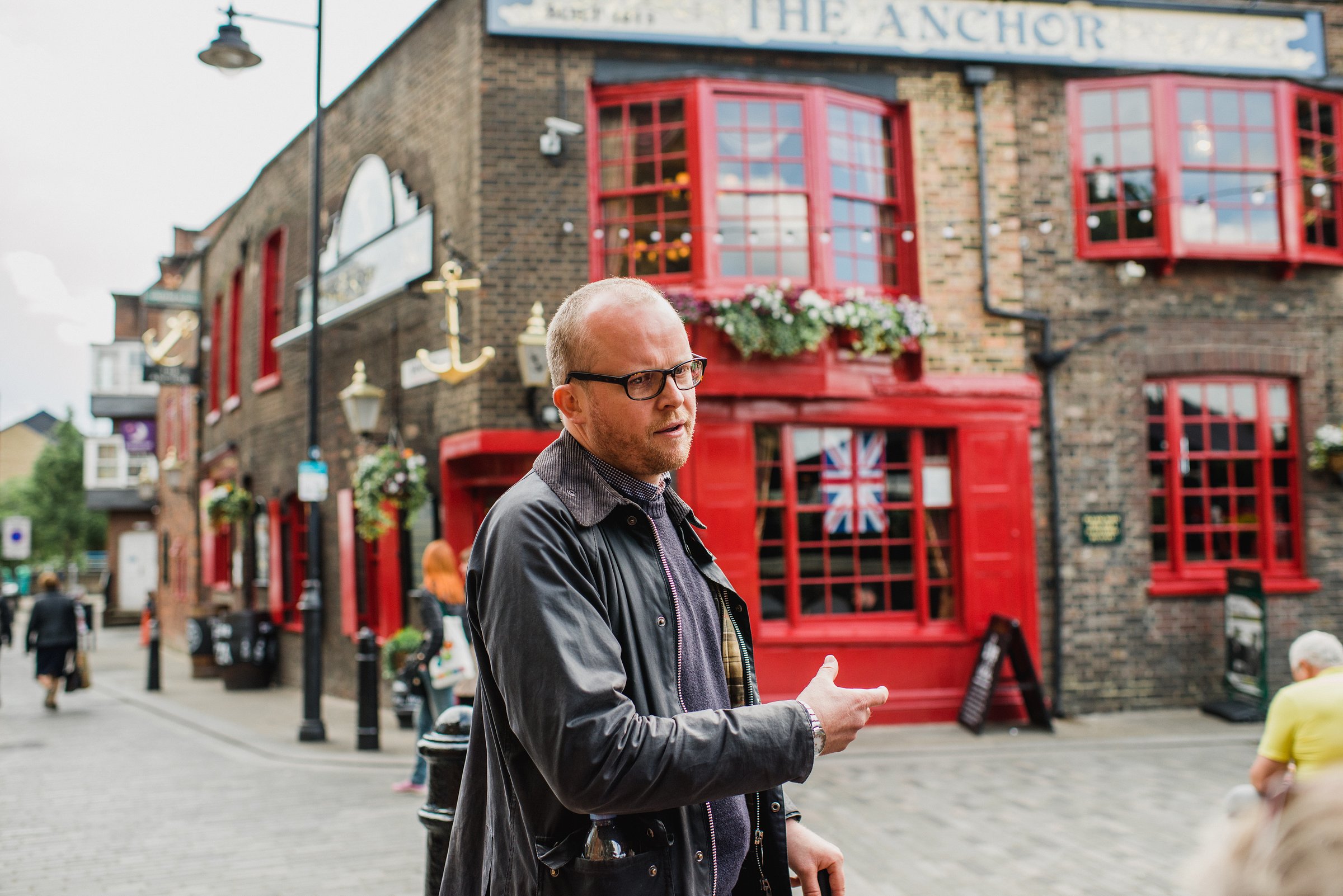 A guide gestures while leading a tour outside The Anchor pub with Union Jack in the window.