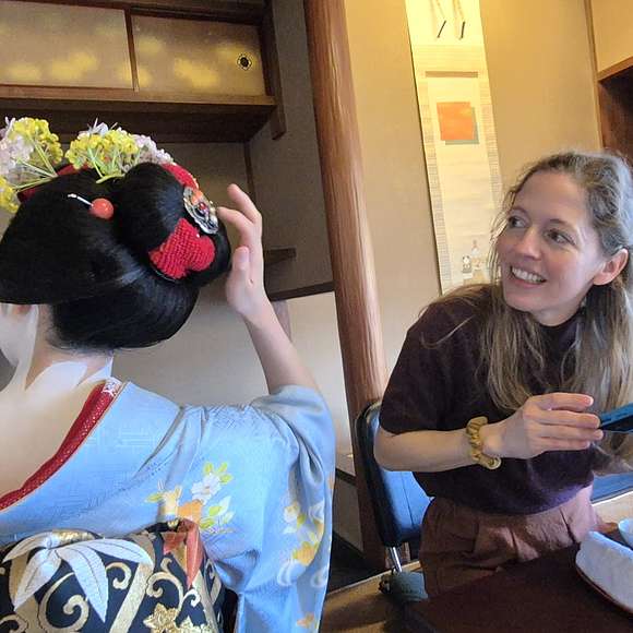 A smiling tourist sits across from a geisha in a blue kimono and ornate hairstyle inside a traditional Japanese room, observing and interacting closely during a cultural experience.