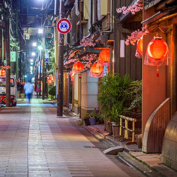 A quiet, lantern-lit alley in Kyoto at night, lined with traditional wooden buildings adorned with red lanterns and artificial cherry blossoms, with a blurred figure walking in the distance.