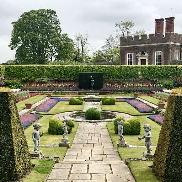 The formal gardens at Hampton Court Palace featuring colorful flower beds, sculpted hedges, small fountains, and stone cherub statues along a central path.