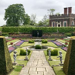 The formal gardens at Hampton Court Palace featuring colorful flower beds, sculpted hedges, small fountains, and stone cherub statues along a central path.