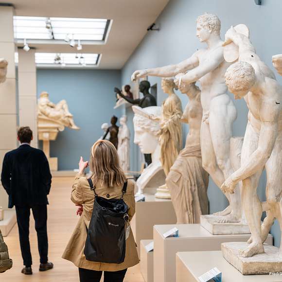 Visitors walk among classical marble statues in a bright exhibition hall.