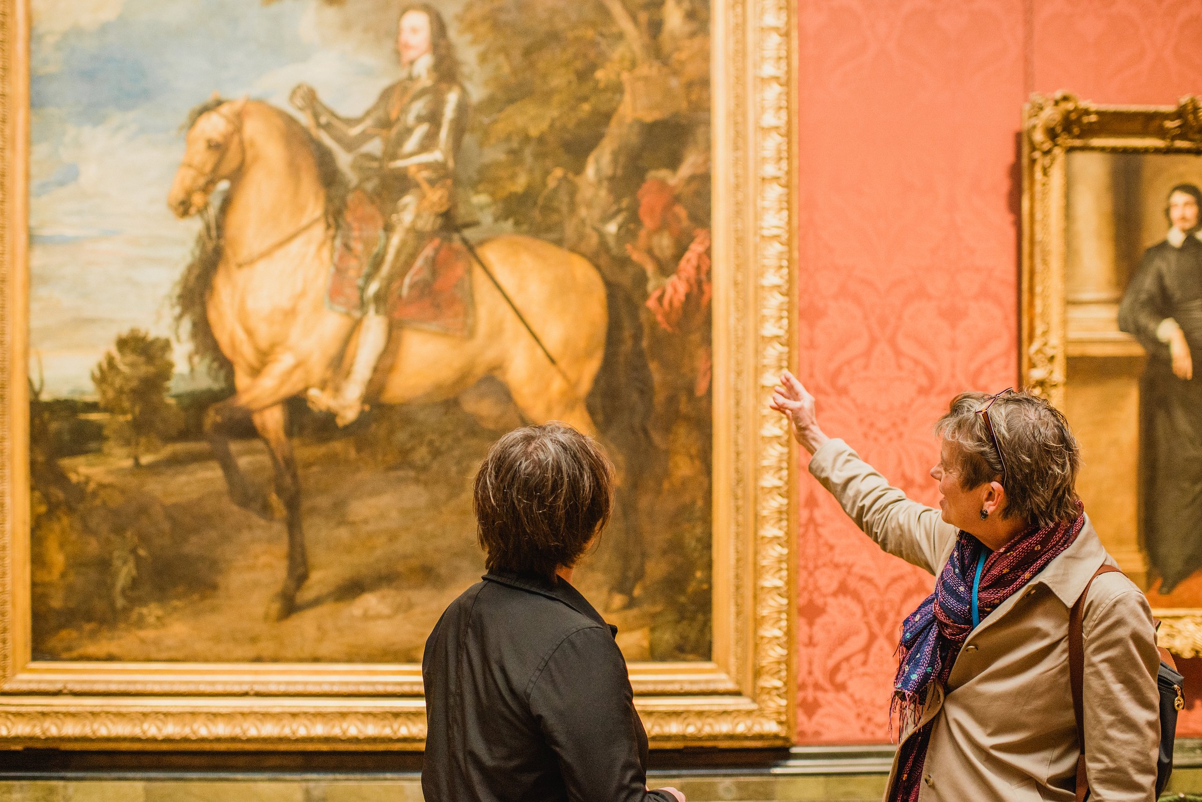 Tour guide gesturing toward a grand equestrian portrait while discussing it with a guest inside the National Gallery.