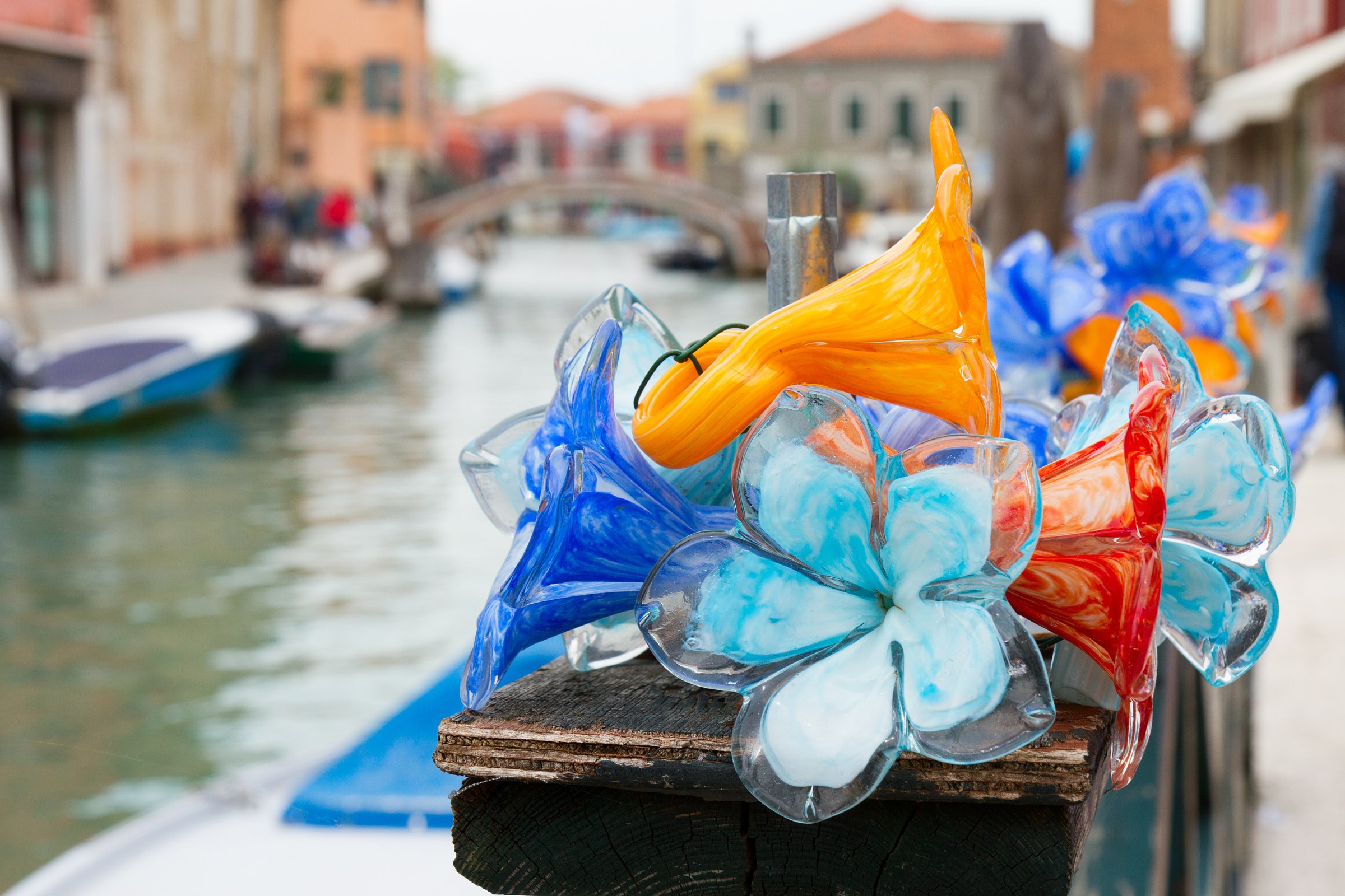 Vibrant Murano glass flowers in blue, orange, and red displayed on a wooden post beside a canal in Venice, with boats and colorful buildings in the background.