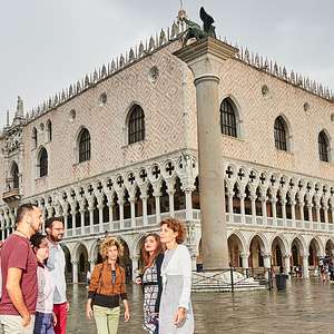 A group of visitors stands outside the Doge's Palace in Venice, admiring its Gothic façade with pink-and-white diamond patterned stonework and arched colonnades.