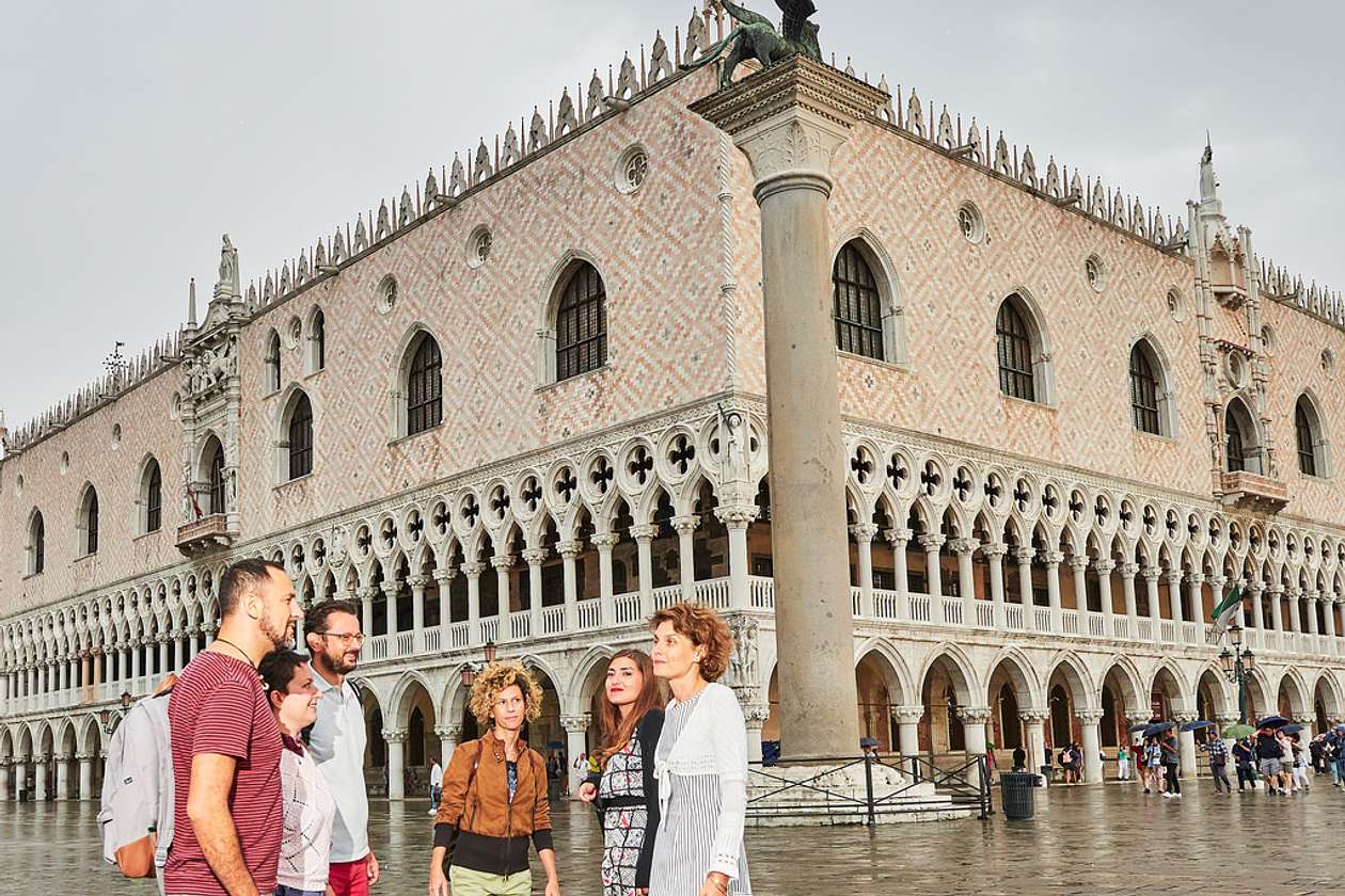 A group of visitors stands outside the Doge's Palace in Venice, admiring its Gothic façade with pink-and-white diamond patterned stonework and arched colonnades.