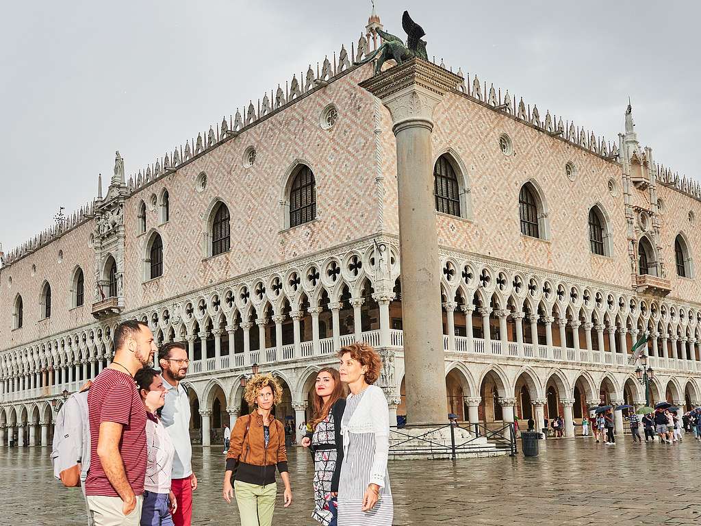 A group of visitors stands outside the Doge's Palace in Venice, admiring its Gothic façade with pink-and-white diamond patterned stonework and arched colonnades.