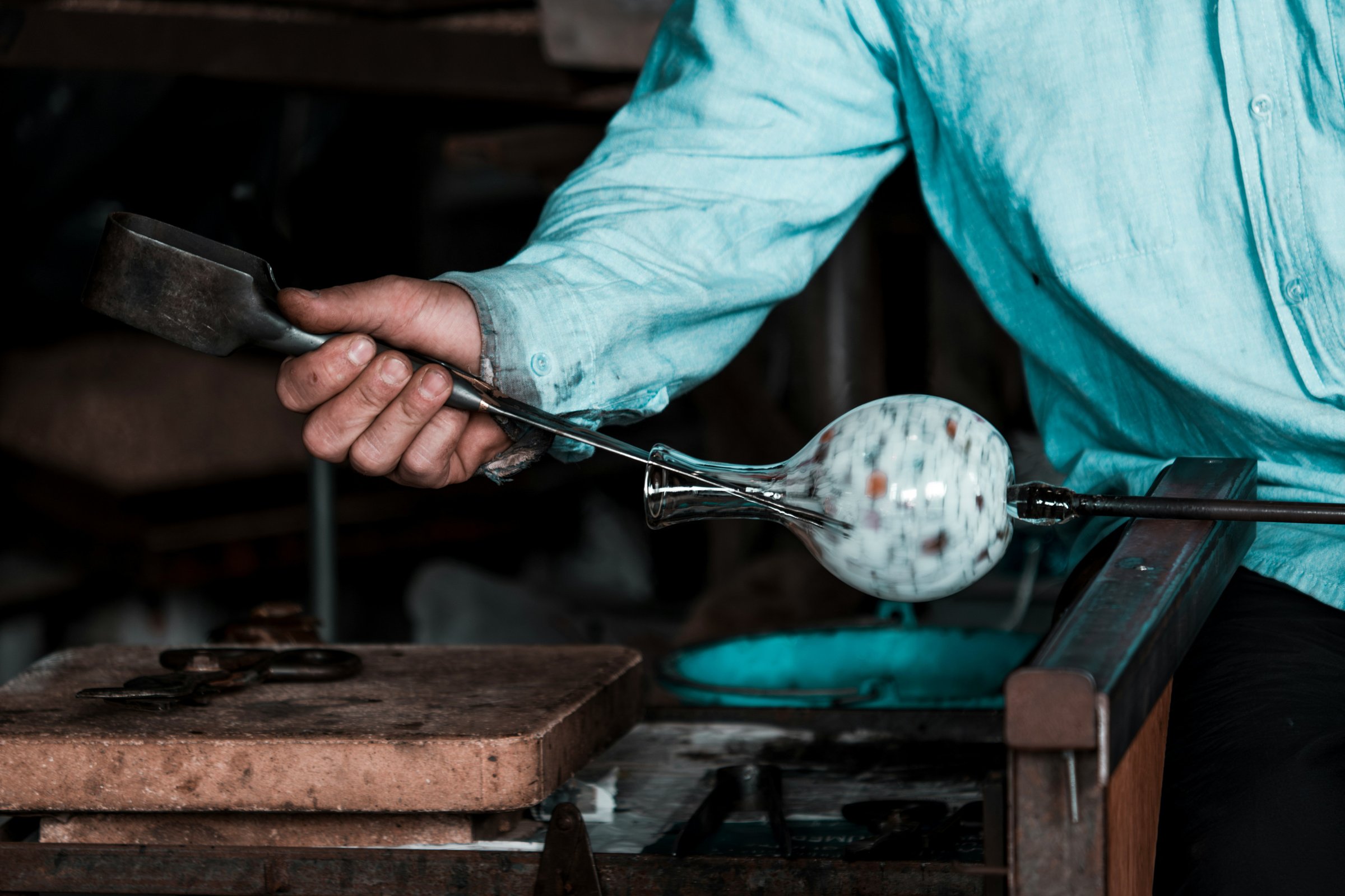 A glassblower in a light blue shirt shapes a speckled glass orb at the end of a blowpipe inside a workshop, using tools to refine its form.