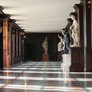 A long wood-paneled gallery inside Hampton Court Palace with marble statues and busts displayed along the walls, sunlight streaming across the checkered floor.
