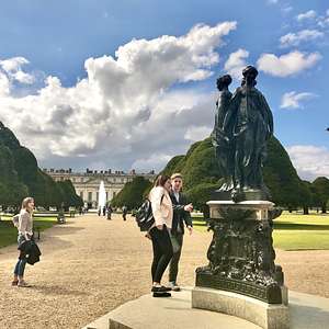 Visitors walking past a bronze statue of two figures in the gardens of Hampton Court Palace, with manicured trees and the palace fountain in the background.