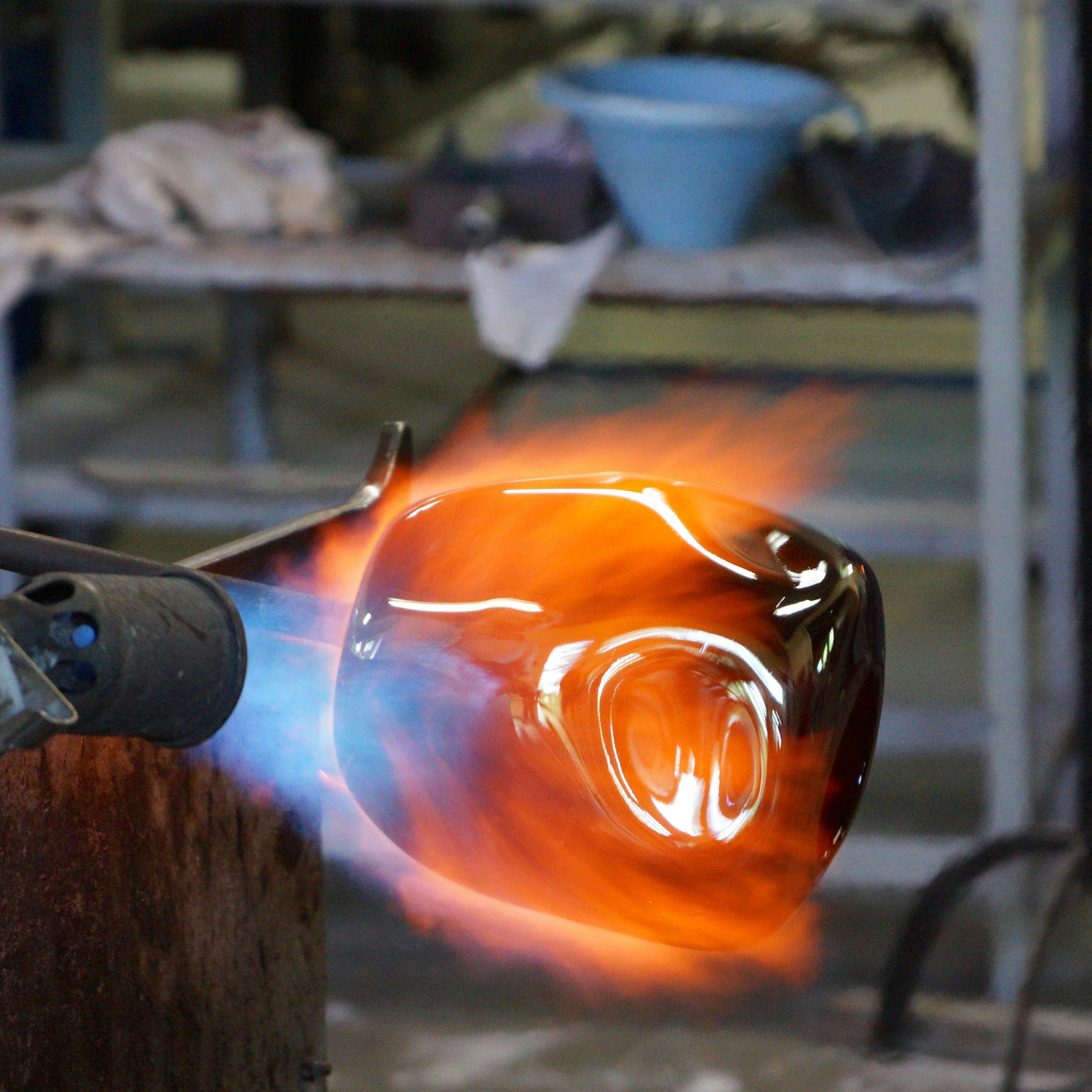 A close-up view of a molten glass object being shaped with a torch in a glassblowing workshop, with vivid orange and blue flames surrounding the glowing glass.