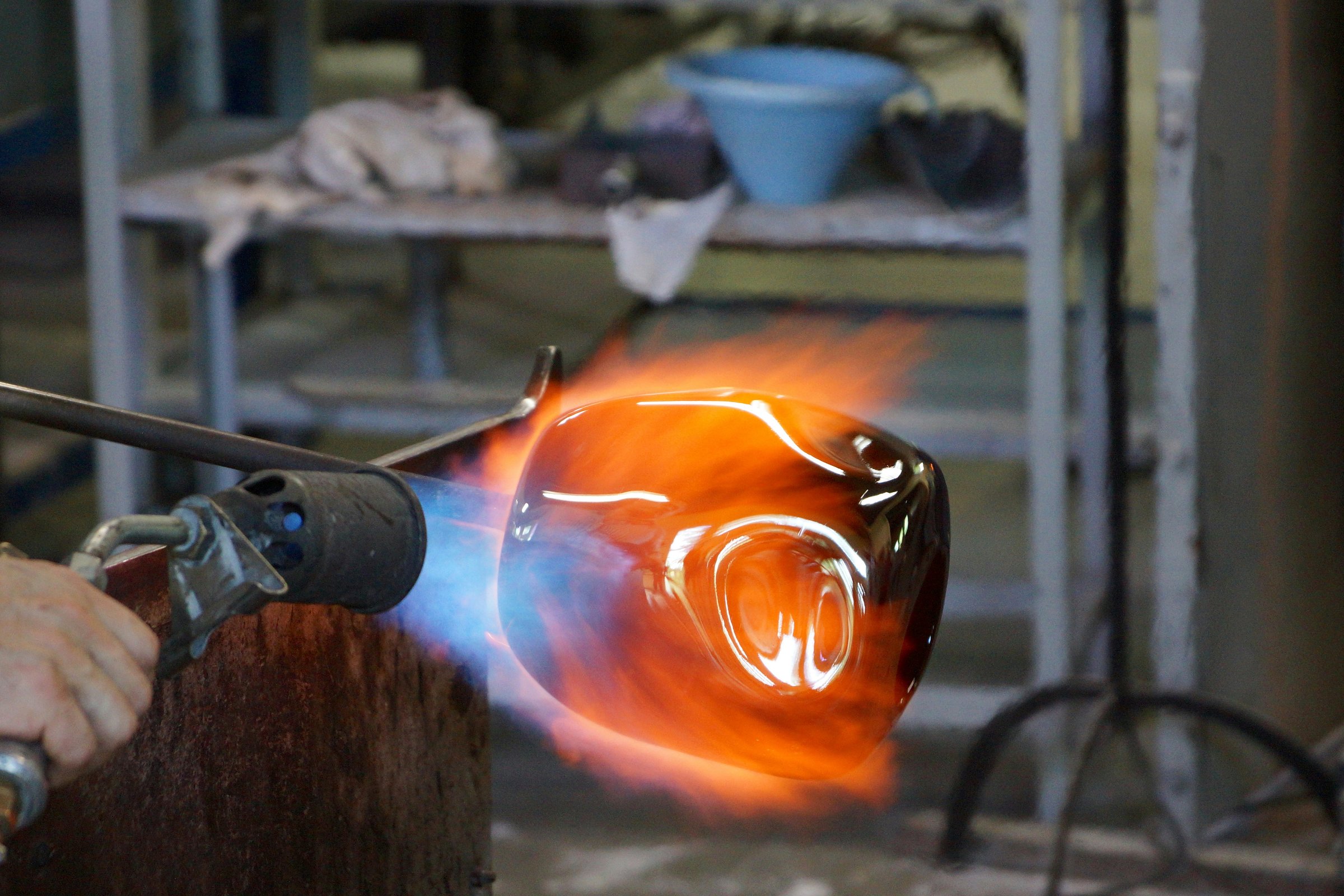 A close-up view of a molten glass object being shaped with a torch in a glassblowing workshop, with vivid orange and blue flames surrounding the glowing glass.