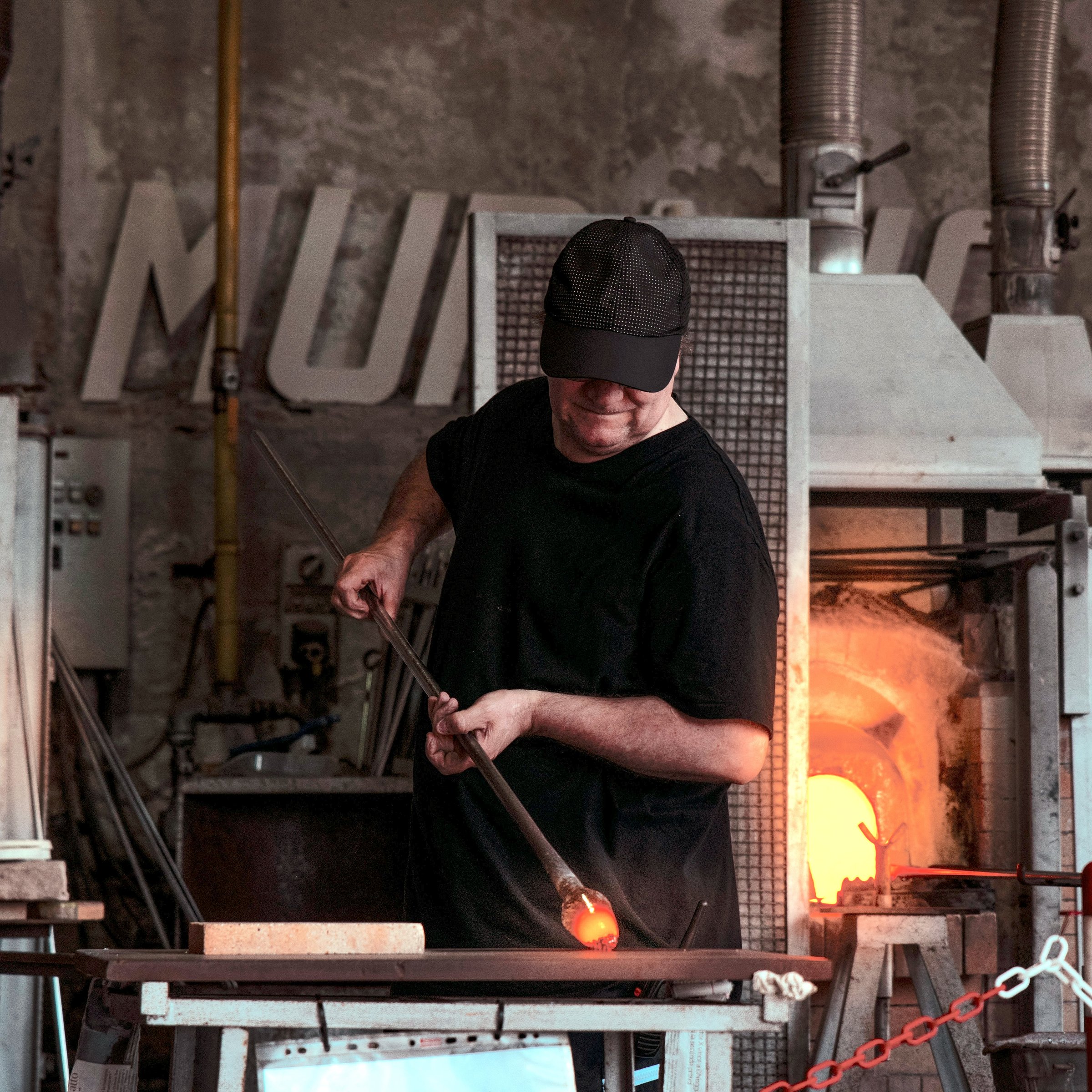 A glassblower wearing a black cap and shirt works with a molten glass piece at the end of a blowpipe in front of a glowing furnace inside a rustic studio.
