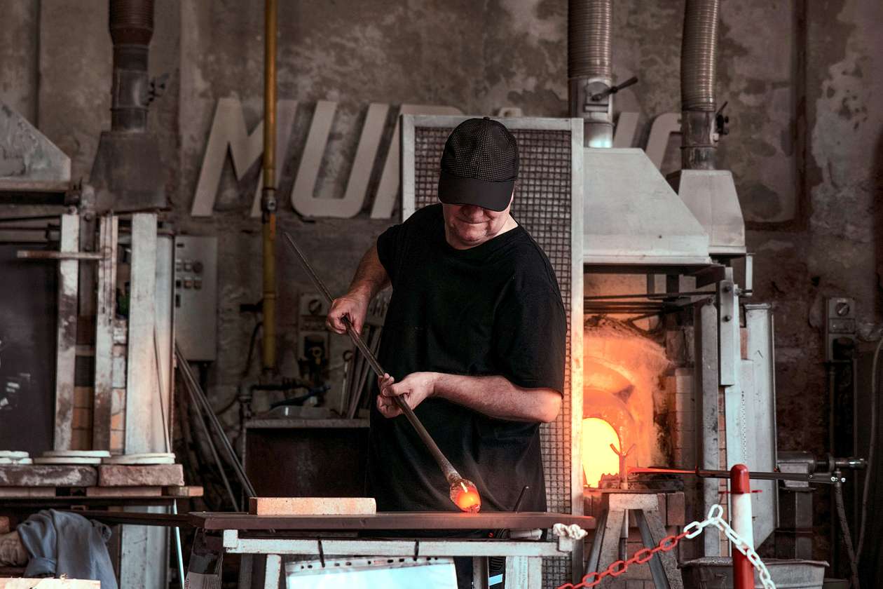 A glassblower wearing a black cap and shirt works with a molten glass piece at the end of a blowpipe in front of a glowing furnace inside a rustic studio.