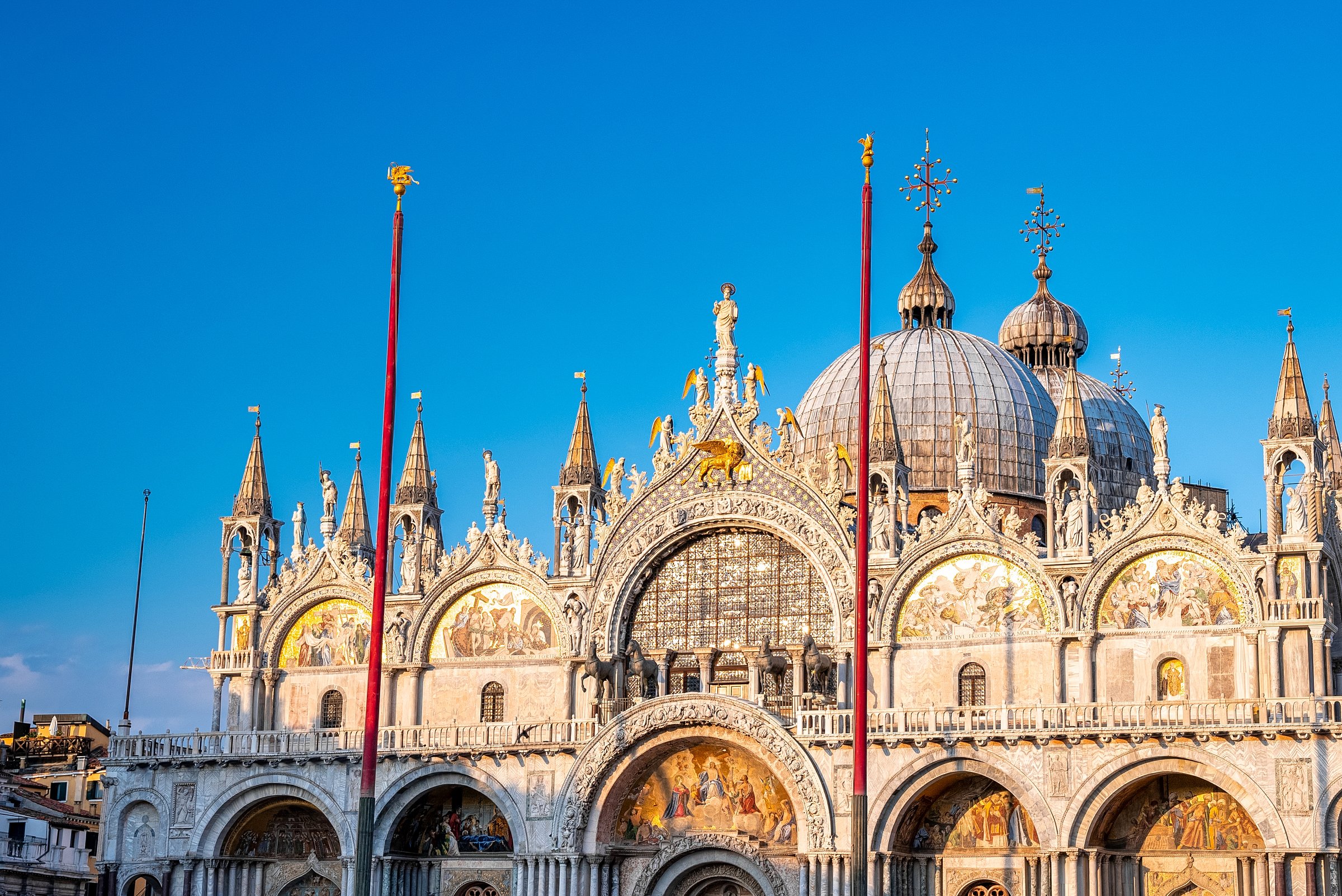St. Mark’s Basilica glowing at golden hour, with domes, mosaics, and statues shining against a blue sky.