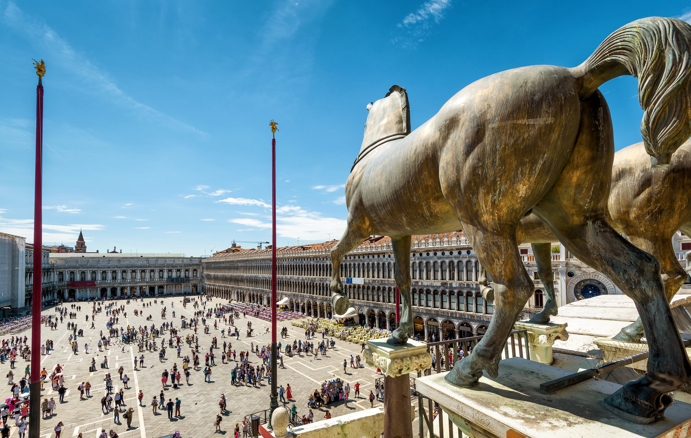 View of Piazza San Marco from the basilica terrace, framed by bronze horse statues and red flagpoles.