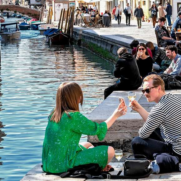 People relax with drinks along a quiet canal in Cannaregio on a sunny afternoon.