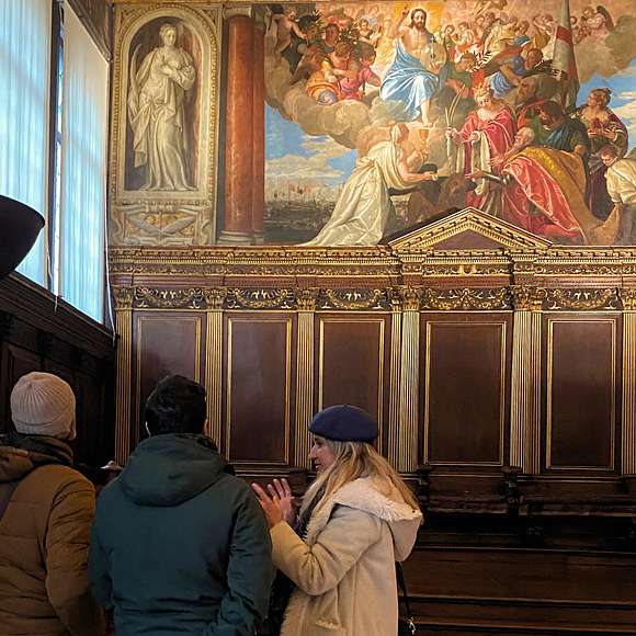 A small group listens to a guide inside the Doge’s Palace, standing before a large Renaissance painting depicting a religious and ceremonial scene surrounded by ornate wood paneling.