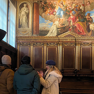 A small group listens to a guide inside the Doge’s Palace, standing before a large Renaissance painting depicting a religious and ceremonial scene surrounded by ornate wood paneling.