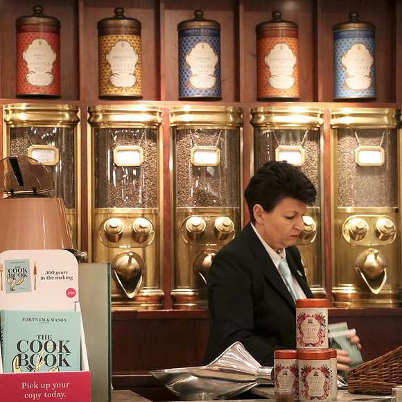 Staff member at a specialty tea and coffee counter in Fortnum & Mason, with decorative tins and grinders behind her.