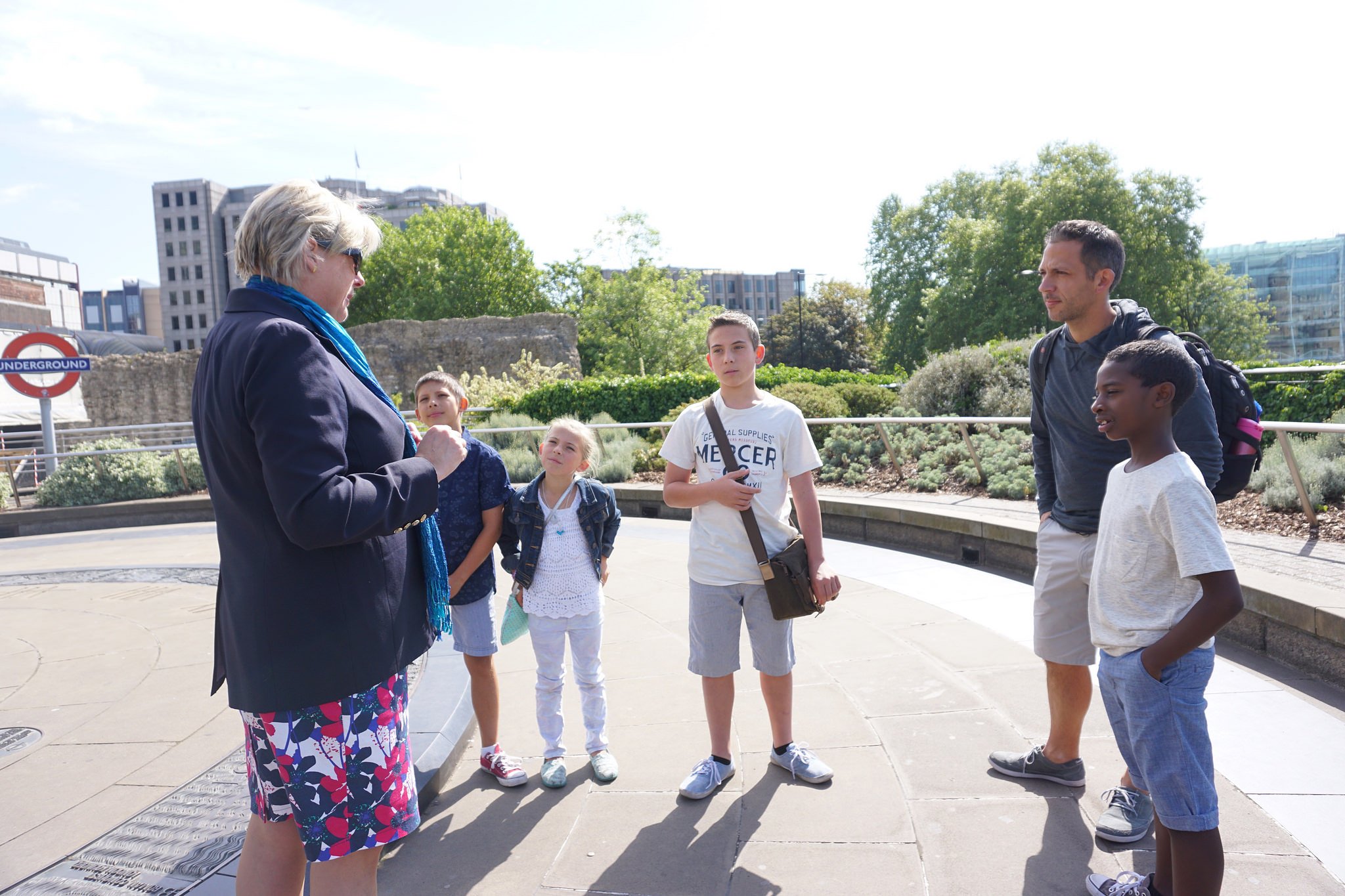 Tour guide engaging a group of children and an adult outside near the Tower of London ruins, with London Underground signage visible.