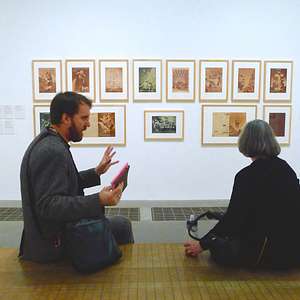 Man and woman seated in discussion at Tate Modern in front of a wall of framed sepia-tone prints.