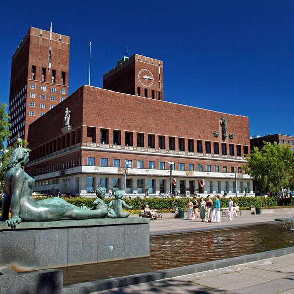Red brick Oslo City Hall with statues and fountains in the foreground under a clear blue sky.