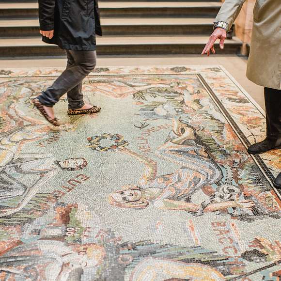 Visitors walking and pointing at a vibrant Roman-style mosaic floor inside the National Gallery, filled with classical figures.