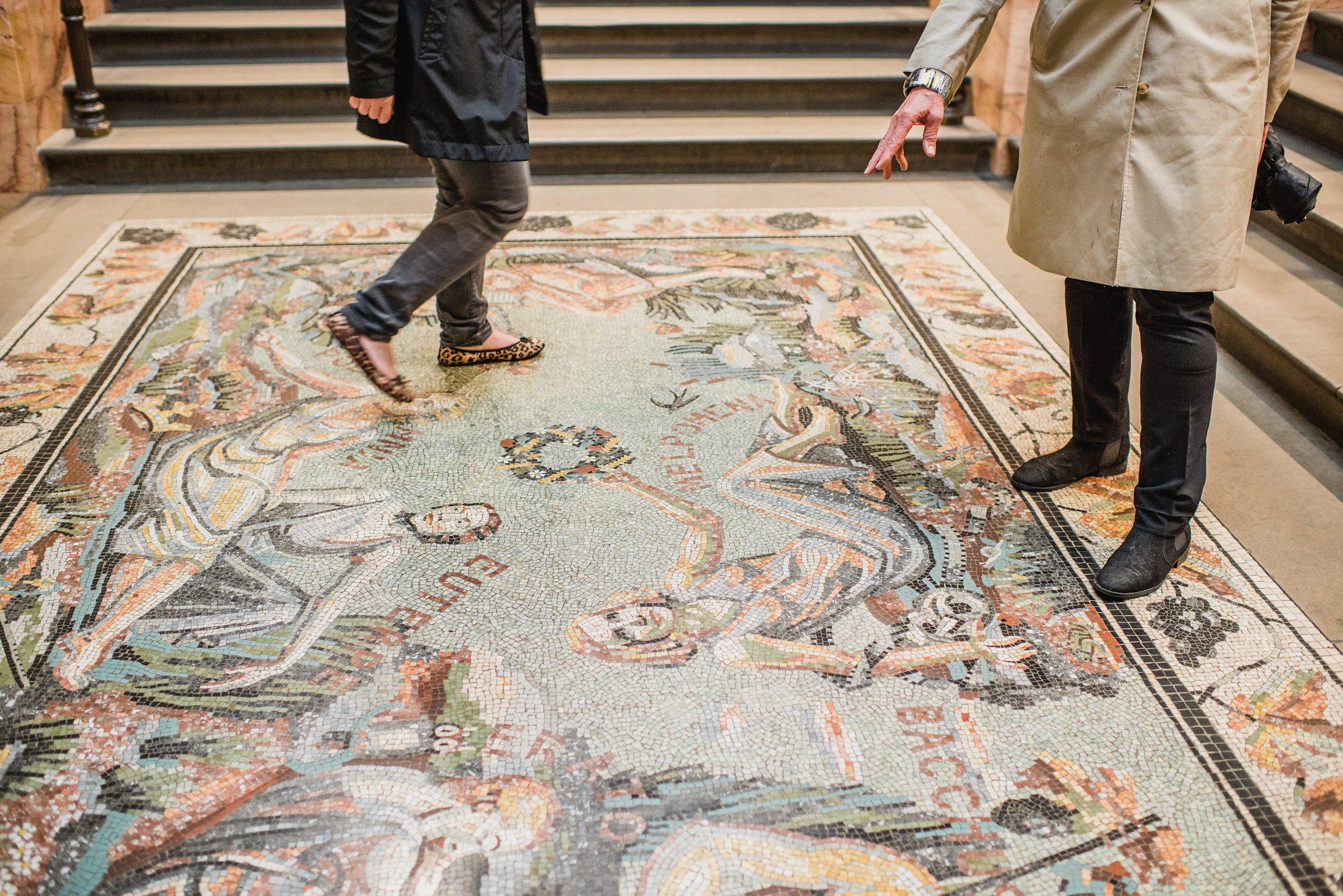 Visitors walking and pointing at a vibrant Roman-style mosaic floor inside the National Gallery, filled with classical figures.