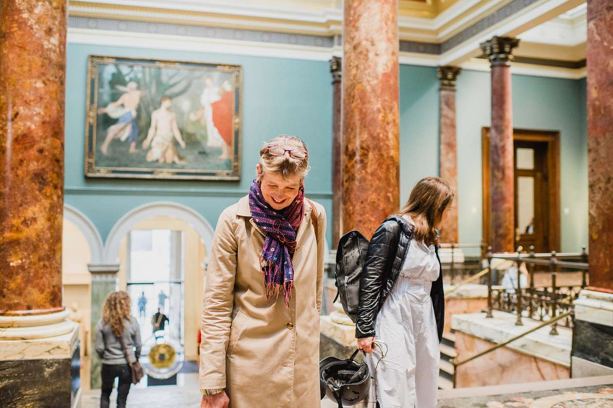 Two women ascending a grand staircase inside the National Gallery, surrounded by marble columns and classical art.
