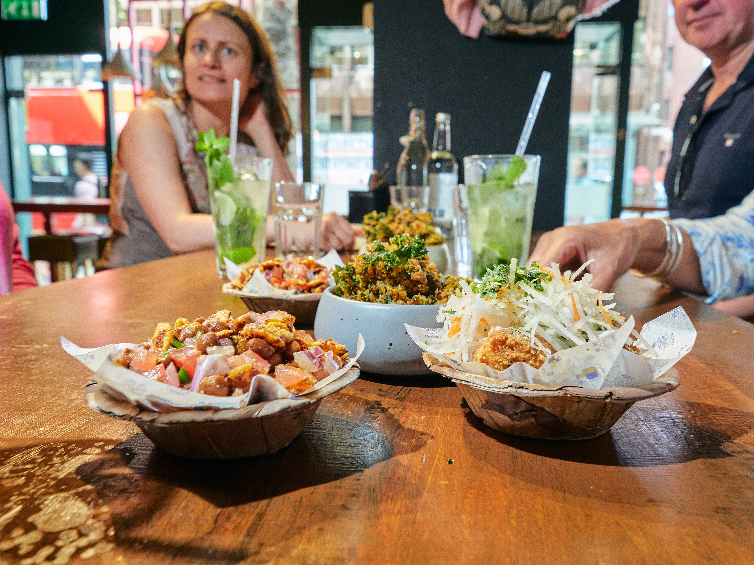 Close-up of colorful dishes on a wooden table with people seated and drinks in hand during a London food tour.