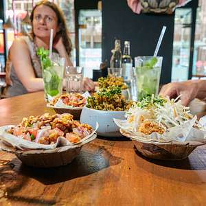 Close-up of colorful dishes on a wooden table with people seated and drinks in hand during a London food tour.